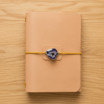A tan leather journal with gold metal rings and a yellow elastic closure wrapped around the front, featuring a decorative blue agate slice charm, set on a wooden tabletop.