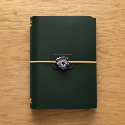 A dark forest green hardcover notebook with a gold elastic closure band and a purple agate slice clasp, laid flat on a wooden surface.