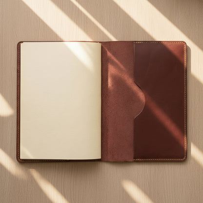 An open leather notebook cover or planner sleeve on a wooden surface, with a blank cream page on the left and a reddish-brown pocket on the right. Sunlight casts diagonal shadows across the scene.