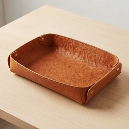A rectangular tan leather tray with a pebbled texture and small metal snap details on the sides, shown on a light wood tabletop.